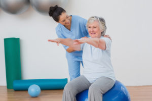 A smiling senior women in physical therapy with her arms raised to the front at shoulder height while sitting on a stability ball with a smiling physical therapist assisting her with stabilization by placing her hands on the senior woman's shoulders.